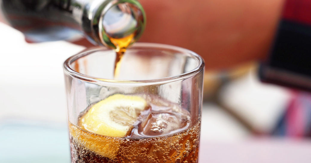 Hand pouring cola soda drink from bottle to glass with ice cubes and lemon slices