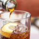 Hand pouring cola soda drink from bottle to glass with ice cubes and lemon slices